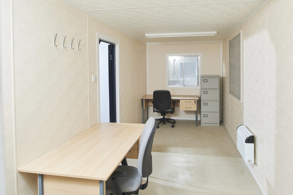 Modern office interior of a steel cabin featuring a desk, chair, filing cabinet, and wall hooks.