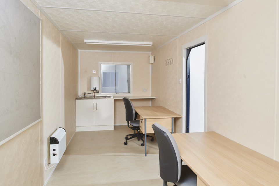 Interior view of a steel office cabin featuring two desks, a kitchenette, and large window.
